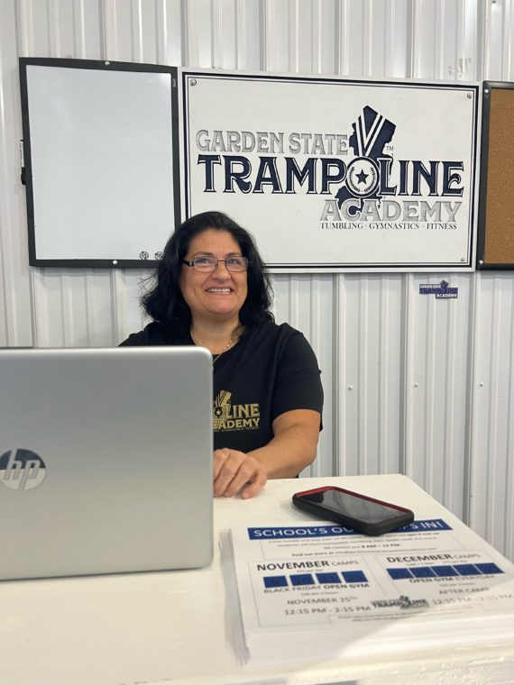 Fran smiling at camera while working on a computer in front of a Garden State Trampoline Academy sign